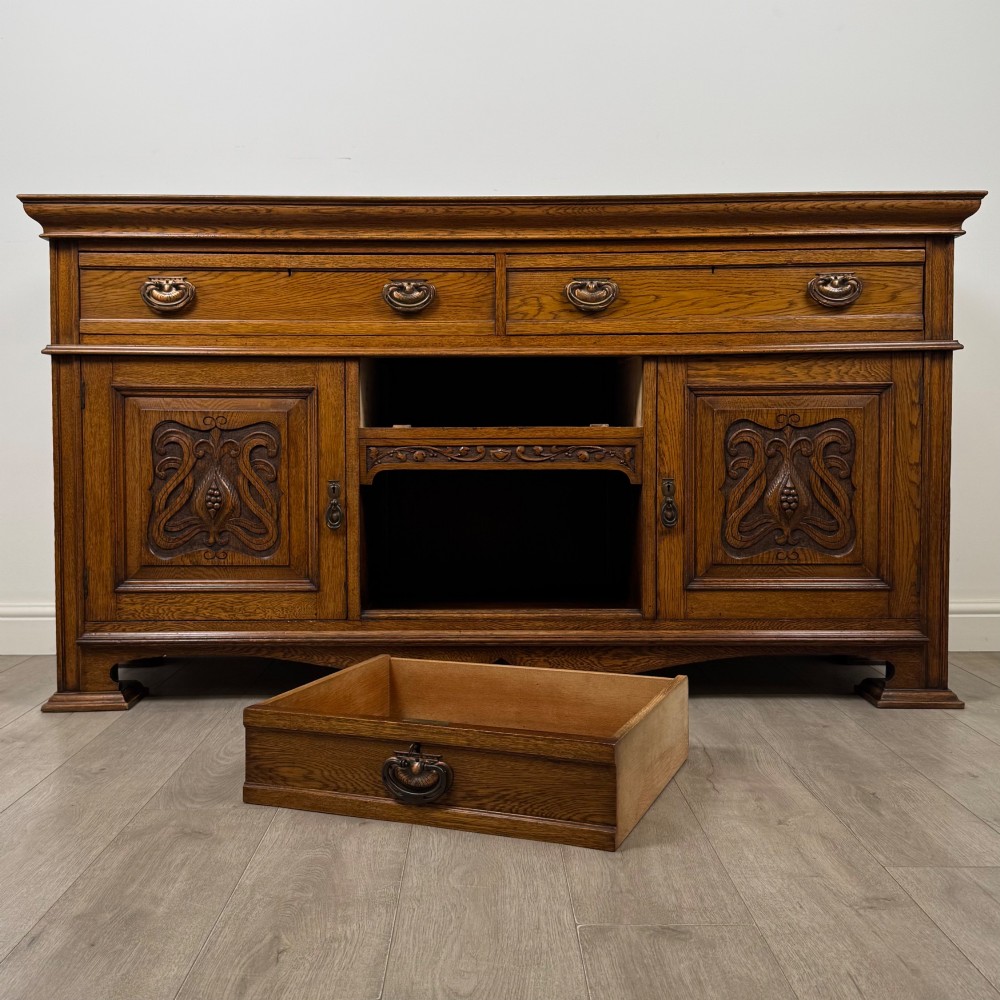 An early 20th-century oak Arts and Crafts sideboard featuring hand-carved foliate panels, copper drop handles, multiple drawers, and a central open recess. View XI