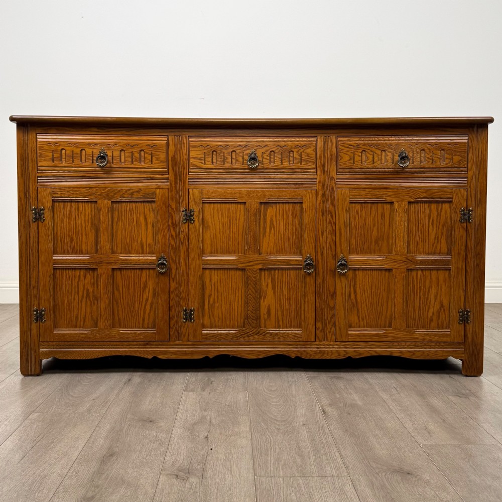 A three-door golden oak sideboard featuring three carved drawers, panelled cabinet doors, and metal drop handles.