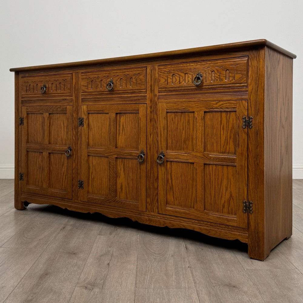 A three-door golden oak sideboard featuring three carved drawers, panelled cabinet doors, and metal drop handles. View III