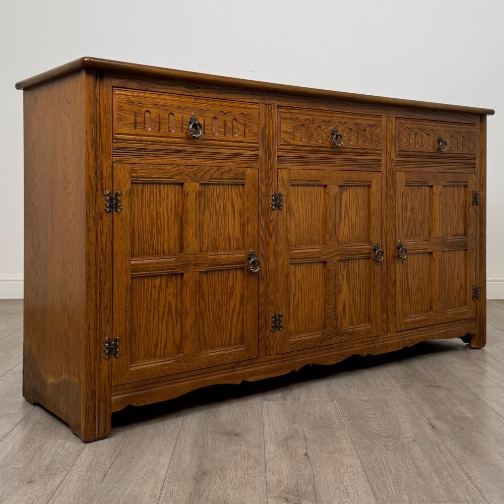 A three-door golden oak sideboard featuring three carved drawers, panelled cabinet doors, and metal drop handles. View IX