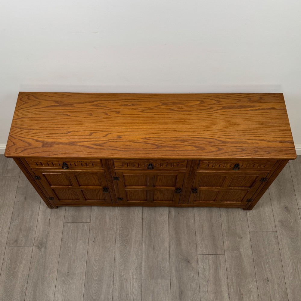 A three-door golden oak sideboard featuring three carved drawers, panelled cabinet doors, and metal drop handles. View X