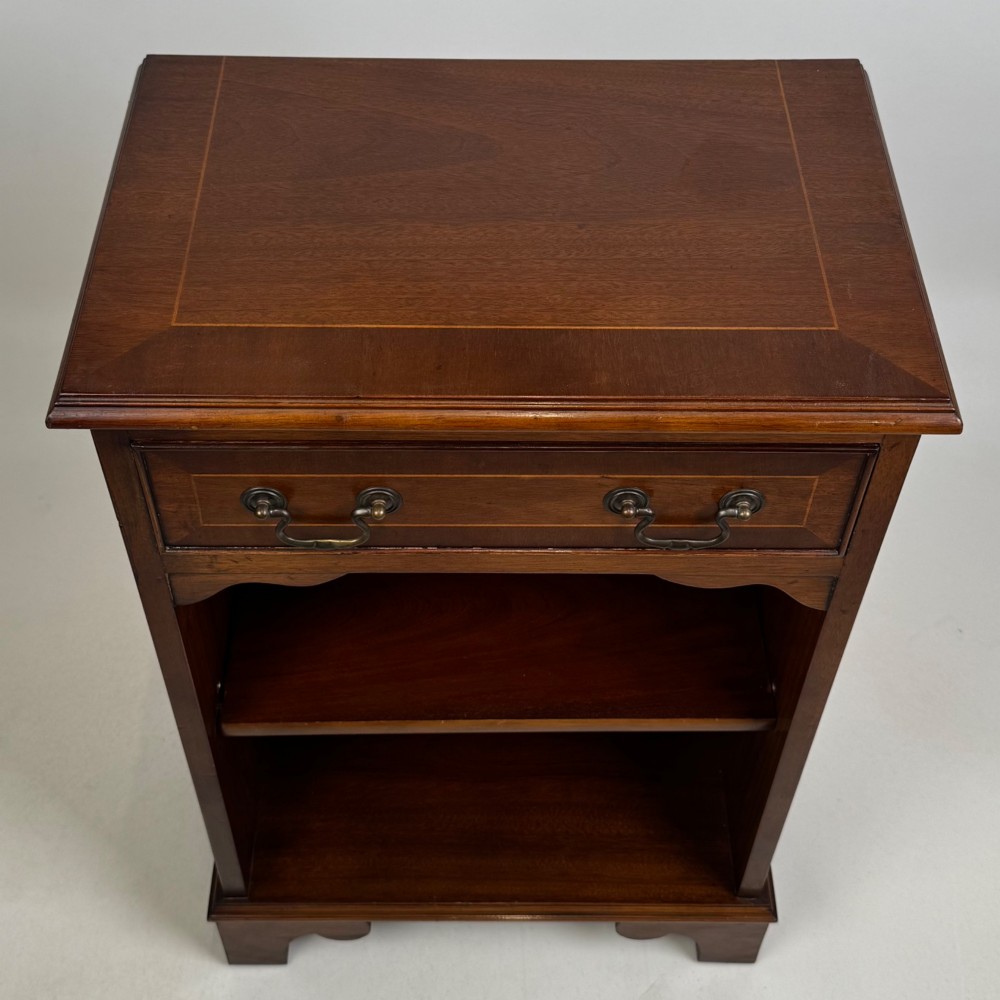 A small mahogany open bookcase featuring a single top drawer with brass swan-neck handles, satinwood inlay, open lower shelving, and shaped bracket feet. View XI