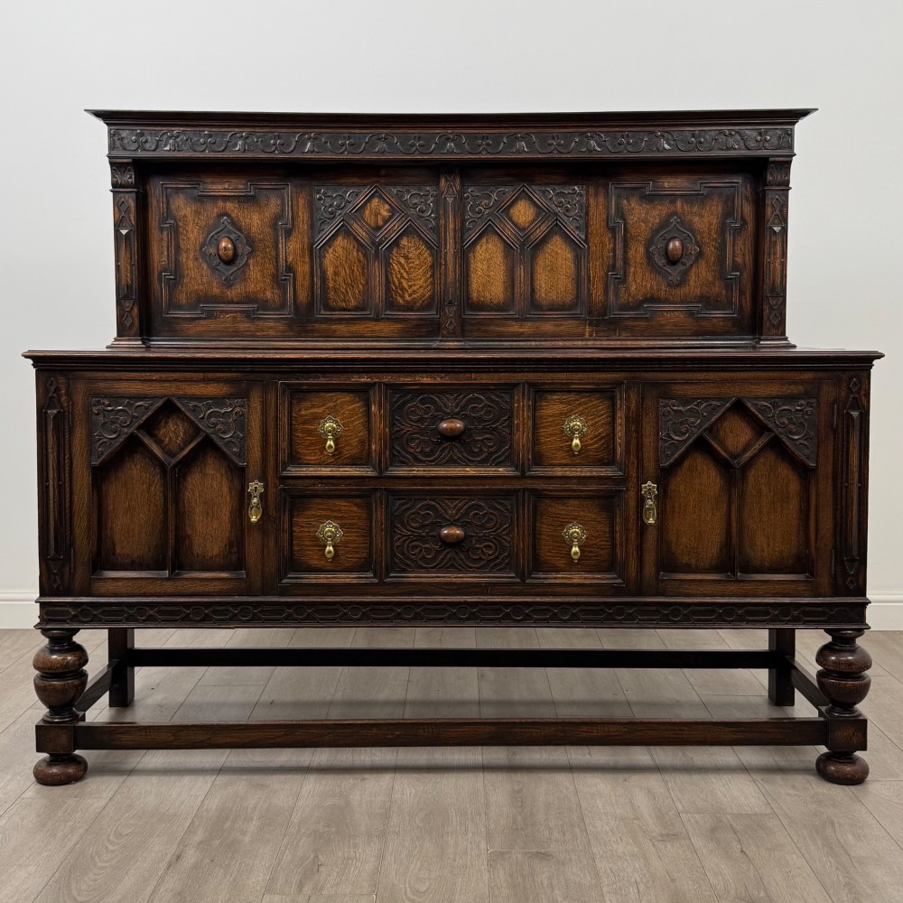 A dark oak Jacobean style sideboard featuring a raised upper superstructure, geometric carved panelling, drawers with brass drop handles, and turned bulbous legs connected by a stretcher base.