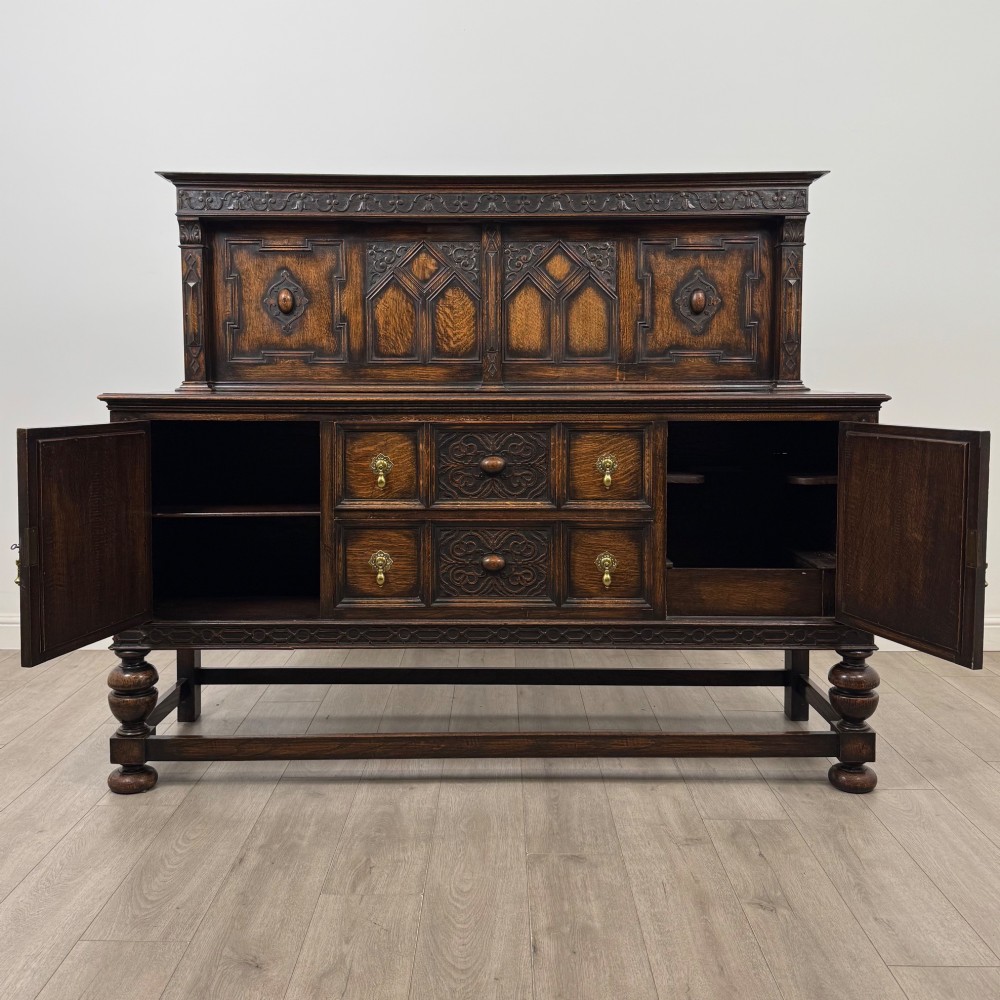 A dark oak Jacobean style sideboard featuring a raised upper superstructure, geometric carved panelling, drawers with brass drop handles, and turned bulbous legs connected by a stretcher base. View II