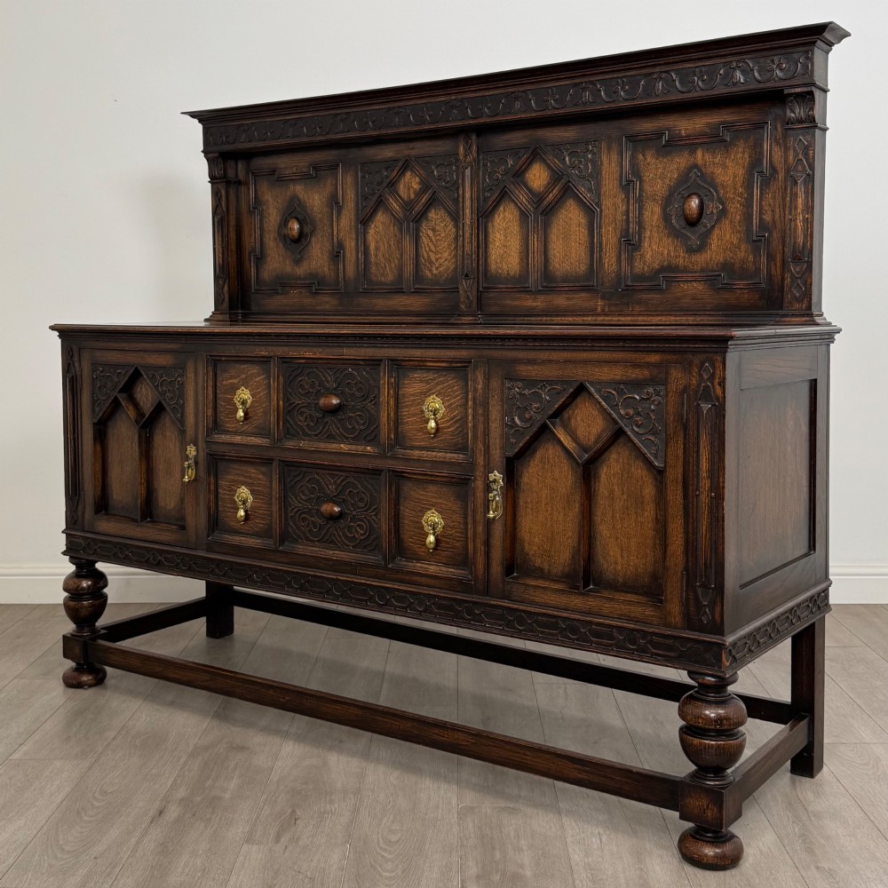 A dark oak Jacobean style sideboard featuring a raised upper superstructure, geometric carved panelling, drawers with brass drop handles, and turned bulbous legs connected by a stretcher base. View III
