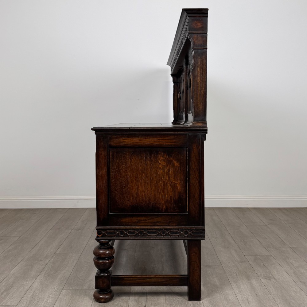 A dark oak Jacobean style sideboard featuring a raised upper superstructure, geometric carved panelling, drawers with brass drop handles, and turned bulbous legs connected by a stretcher base. View IV