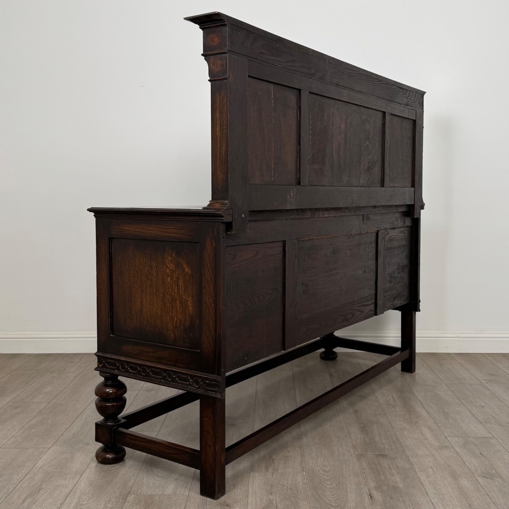 A dark oak Jacobean style sideboard featuring a raised upper superstructure, geometric carved panelling, drawers with brass drop handles, and turned bulbous legs connected by a stretcher base. View V