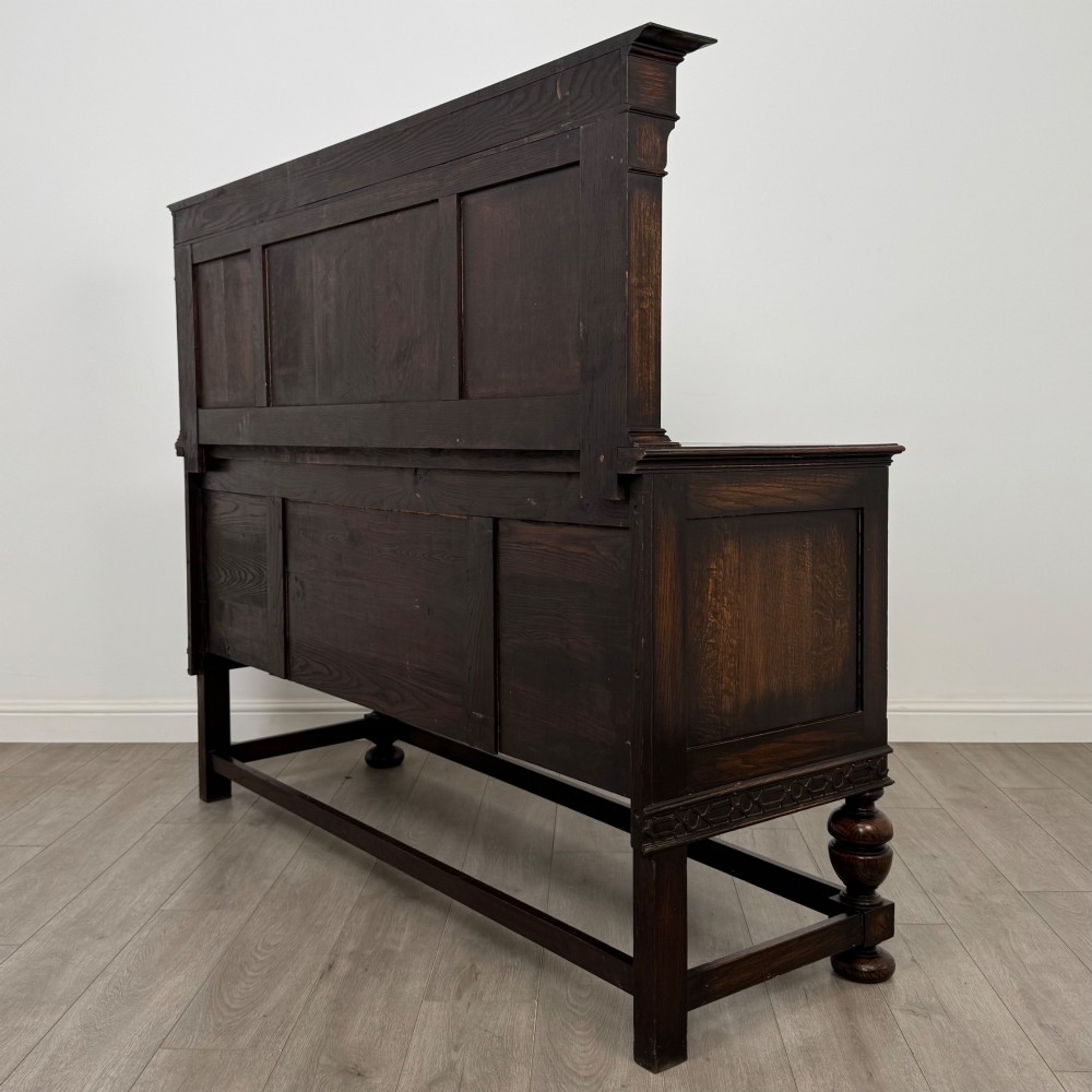 A dark oak Jacobean style sideboard featuring a raised upper superstructure, geometric carved panelling, drawers with brass drop handles, and turned bulbous legs connected by a stretcher base. View VII