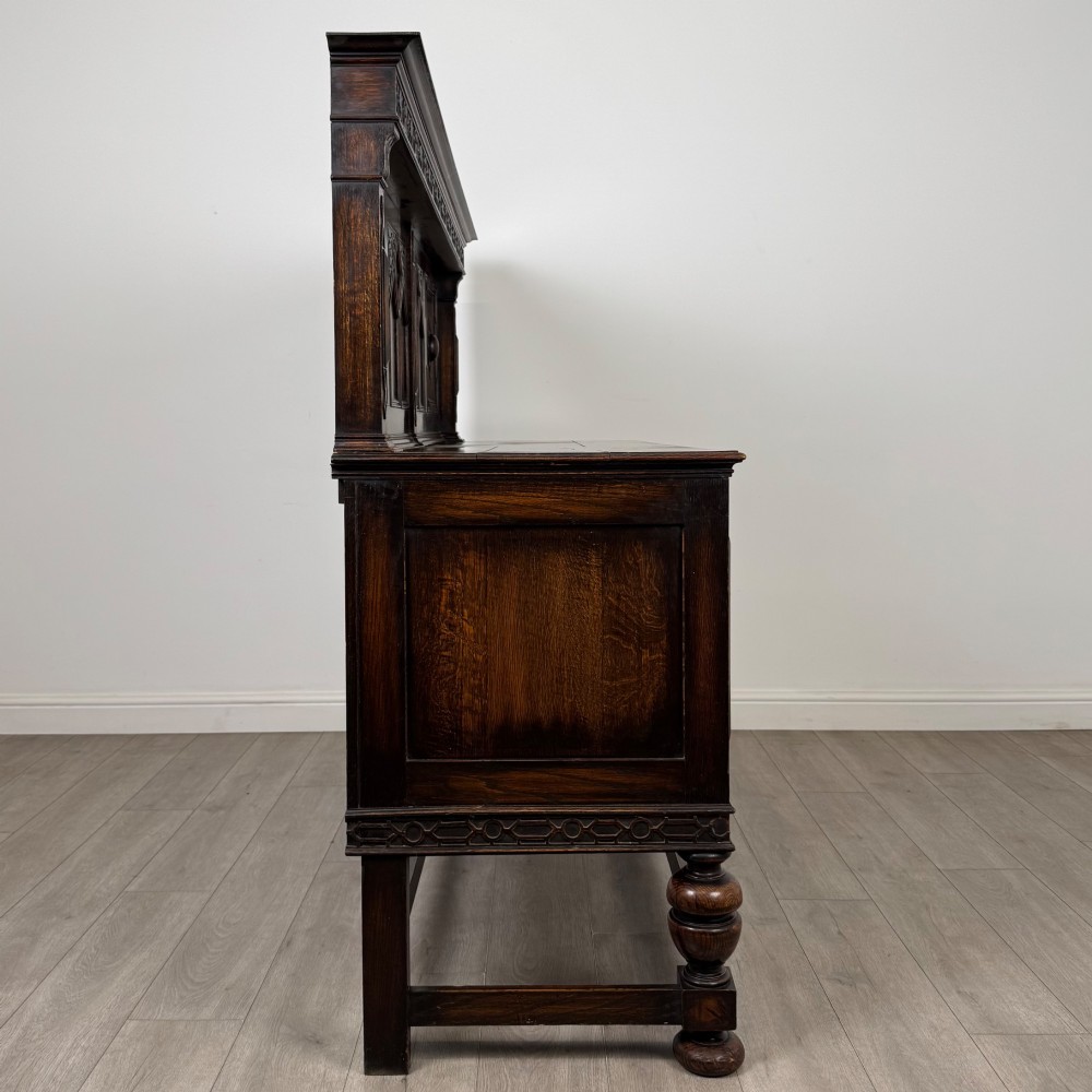 A dark oak Jacobean style sideboard featuring a raised upper superstructure, geometric carved panelling, drawers with brass drop handles, and turned bulbous legs connected by a stretcher base. View VIII