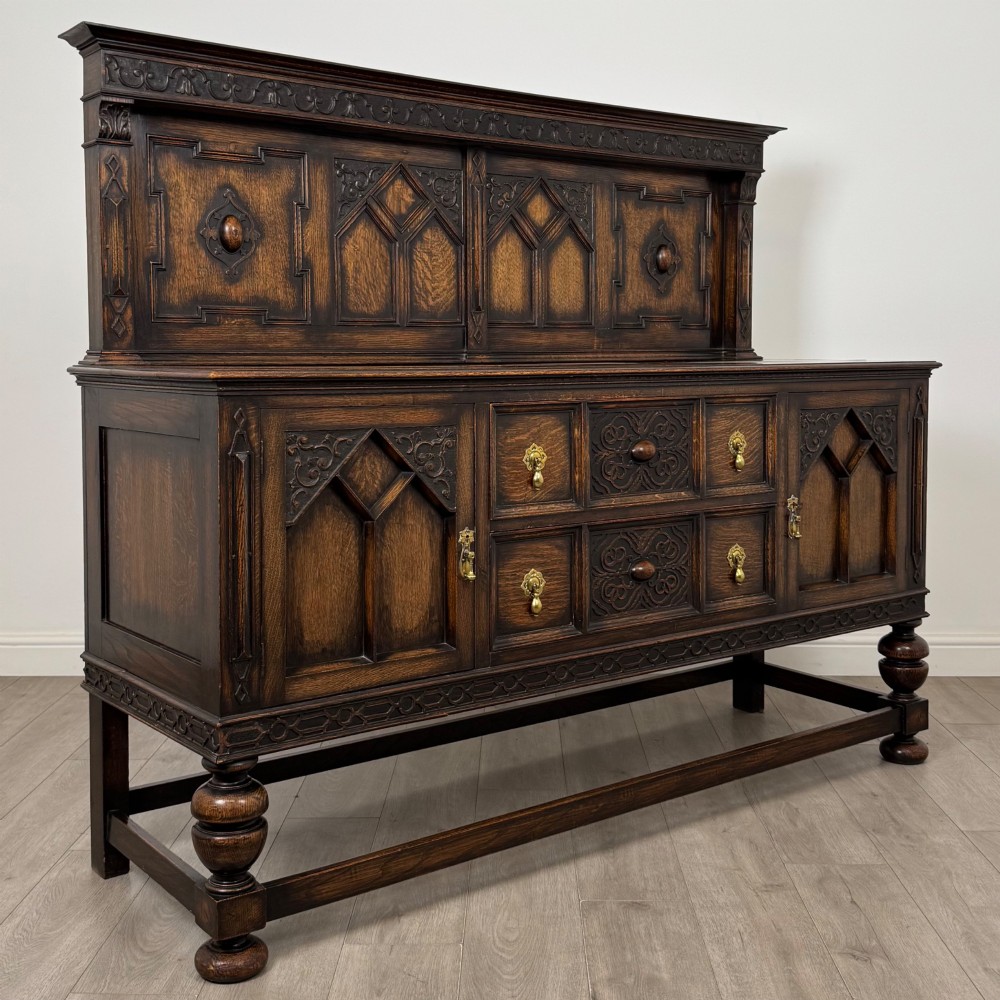A dark oak Jacobean style sideboard featuring a raised upper superstructure, geometric carved panelling, drawers with brass drop handles, and turned bulbous legs connected by a stretcher base. View IX