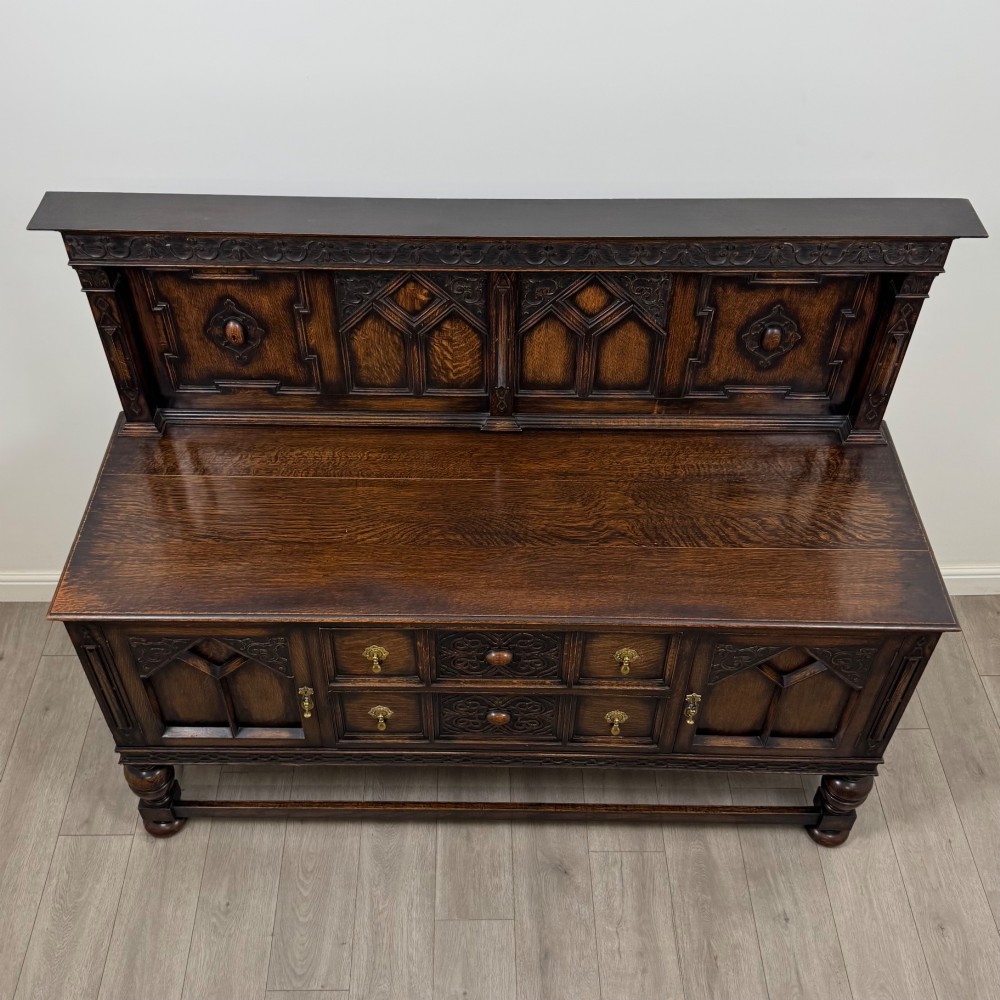 A dark oak Jacobean style sideboard featuring a raised upper superstructure, geometric carved panelling, drawers with brass drop handles, and turned bulbous legs connected by a stretcher base. View X