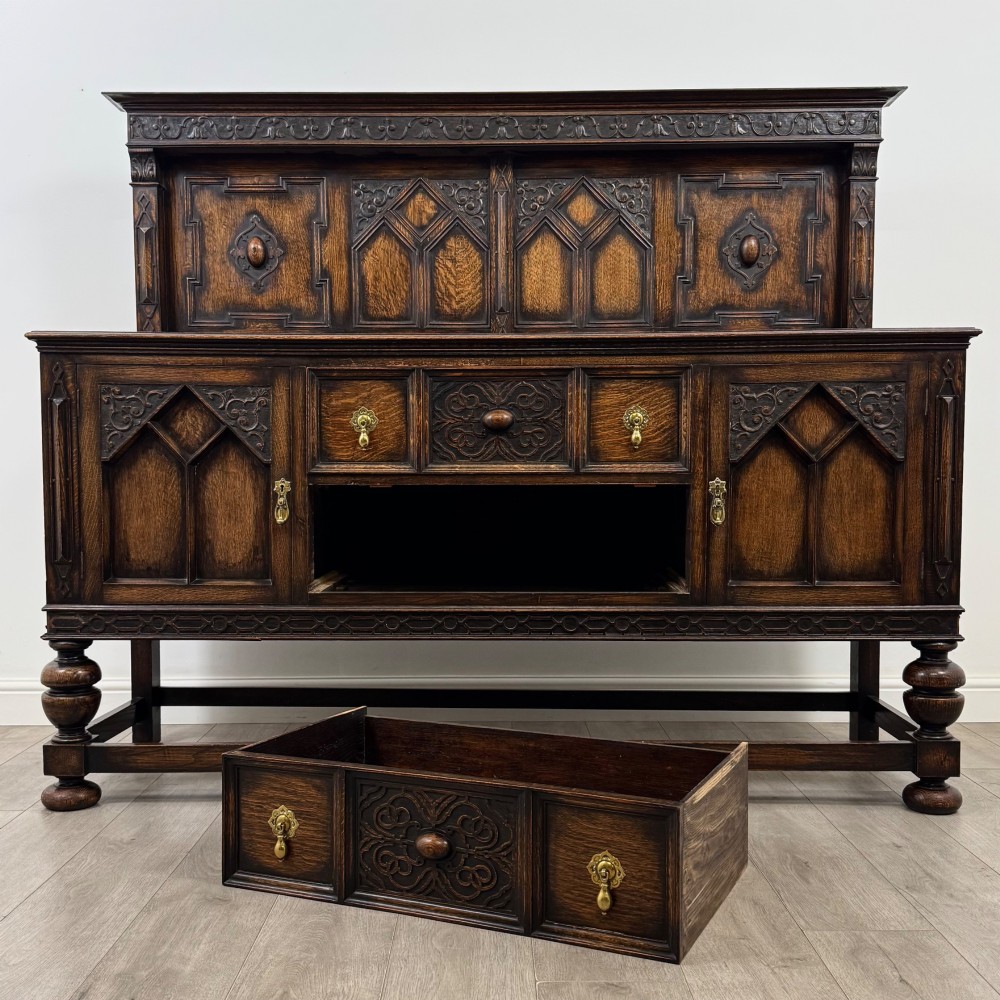 A dark oak Jacobean style sideboard featuring a raised upper superstructure, geometric carved panelling, drawers with brass drop handles, and turned bulbous legs connected by a stretcher base. View XI