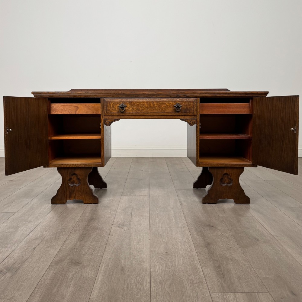 A 20th-century carved oak pedestal desk featuring linenfold cupboard doors, a central top drawer, and trefoil pierced end supports. View II