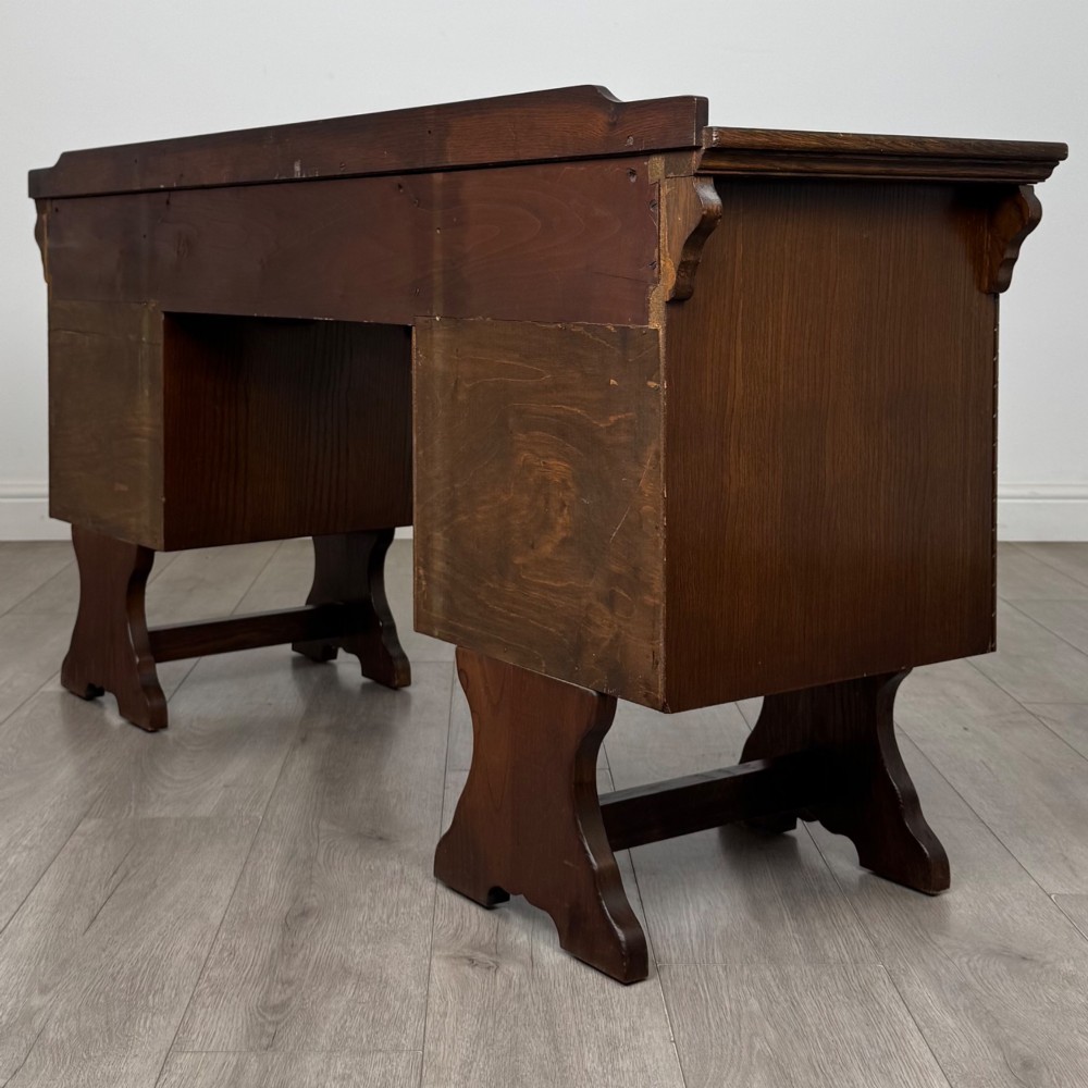 A 20th-century carved oak pedestal desk featuring linenfold cupboard doors, a central top drawer, and trefoil pierced end supports. View VII