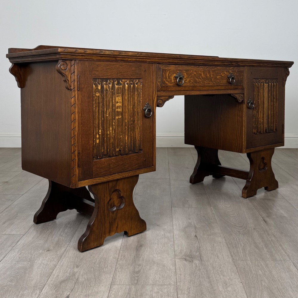 A 20th-century carved oak pedestal desk featuring linenfold cupboard doors, a central top drawer, and trefoil pierced end supports. View IX