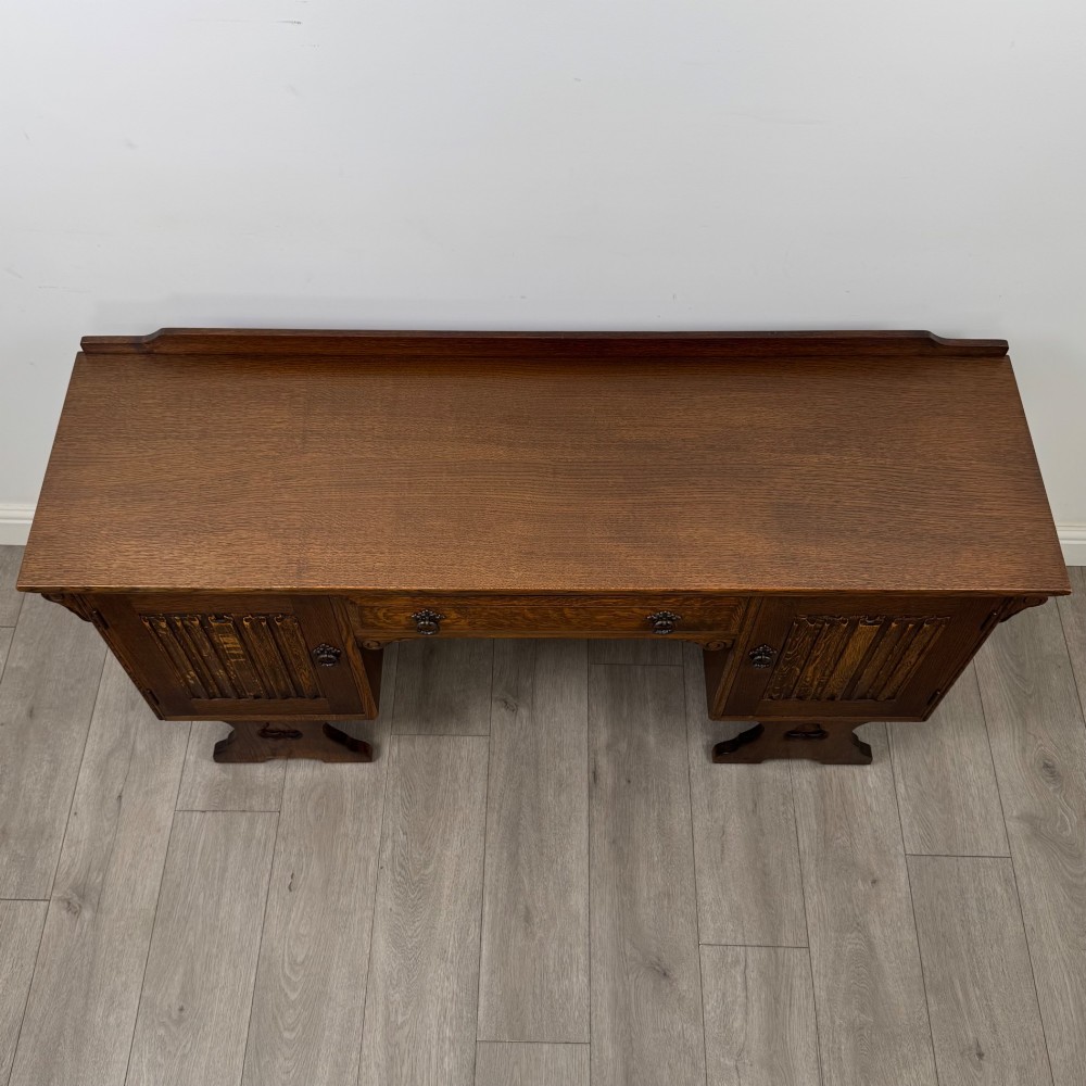 A 20th-century carved oak pedestal desk featuring linenfold cupboard doors, a central top drawer, and trefoil pierced end supports. View X