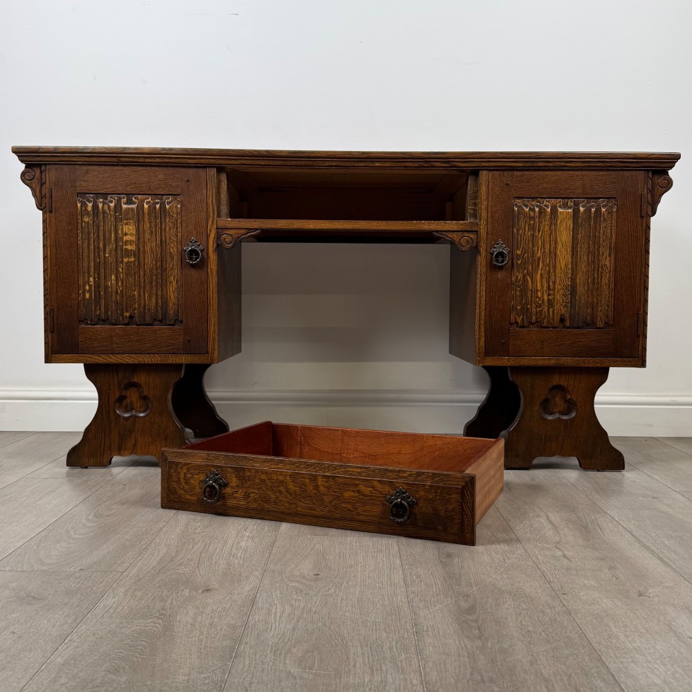 A 20th-century carved oak pedestal desk featuring linenfold cupboard doors, a central top drawer, and trefoil pierced end supports. View XI