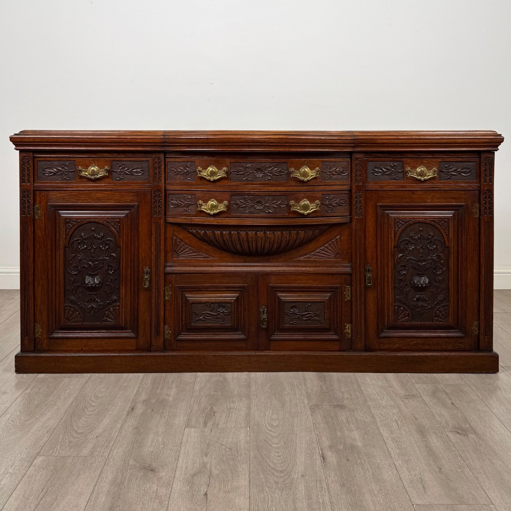 A wide 19th-century Victorian dark oak sideboard featuring relief-carved floral panels, central drawers with brass handles, and lower cupboard doors.