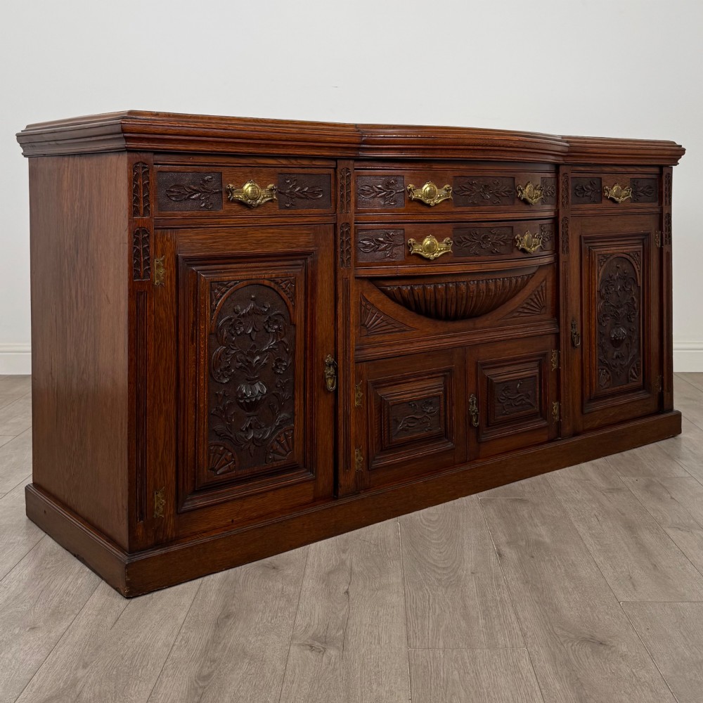 A wide 19th-century Victorian dark oak sideboard featuring relief-carved floral panels, central drawers with brass handles, and lower cupboard doors. View IX