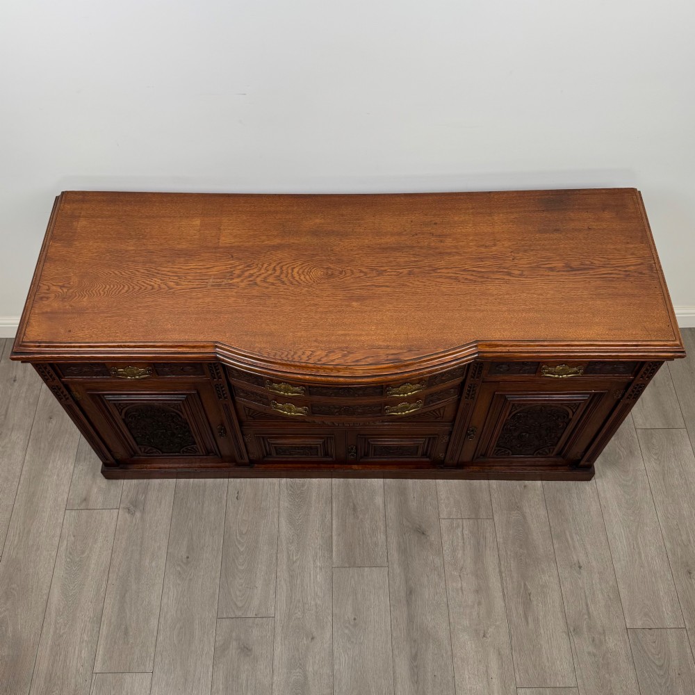 A wide 19th-century Victorian dark oak sideboard featuring relief-carved floral panels, central drawers with brass handles, and lower cupboard doors. View X