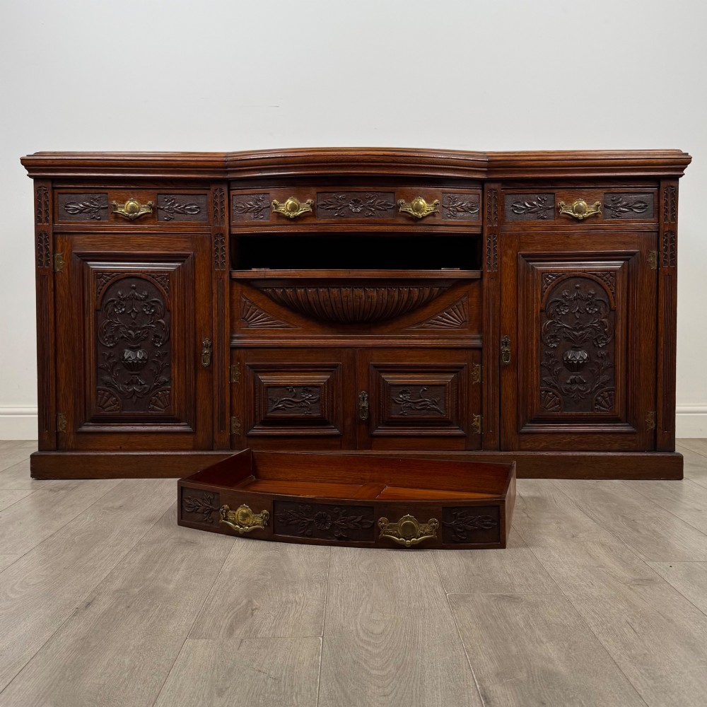 A wide 19th-century Victorian dark oak sideboard featuring relief-carved floral panels, central drawers with brass handles, and lower cupboard doors. View XI