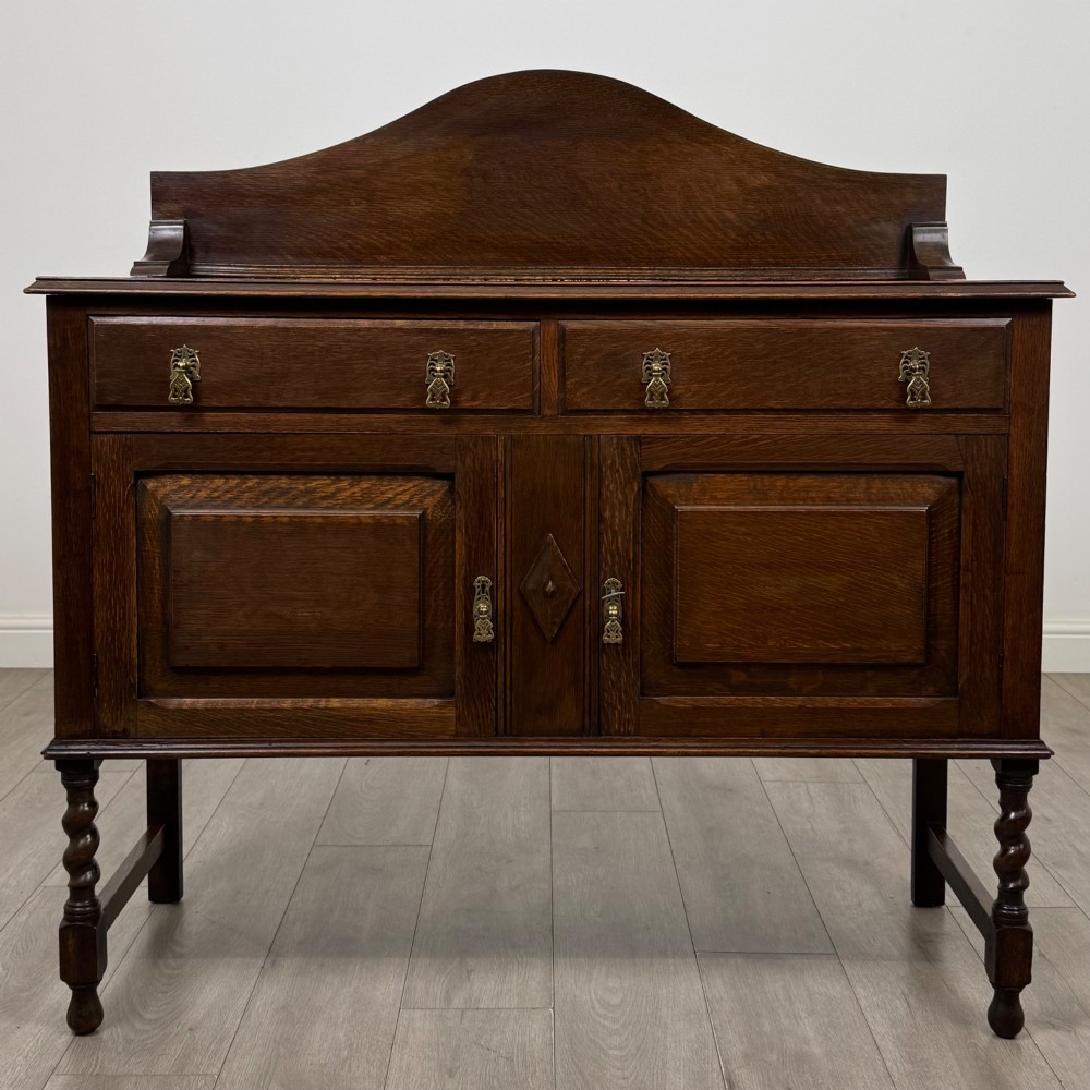 An Edwardian dark oak sideboard featuring a shaped upstand gallery, two top drawers with brass drop handles, twin raised-panel cupboard doors, and front barley twist legs.
