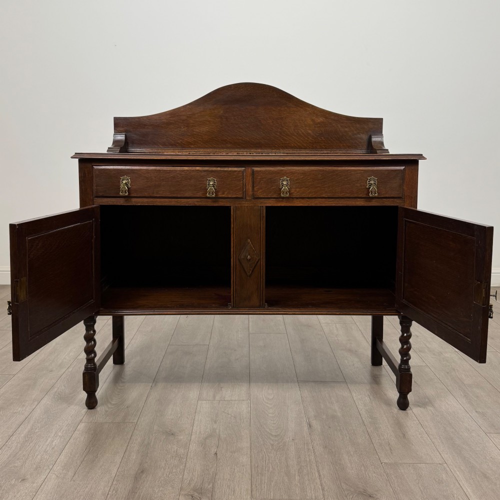 An Edwardian dark oak sideboard featuring a shaped upstand gallery, two top drawers with brass drop handles, twin raised-panel cupboard doors, and front barley twist legs. View II