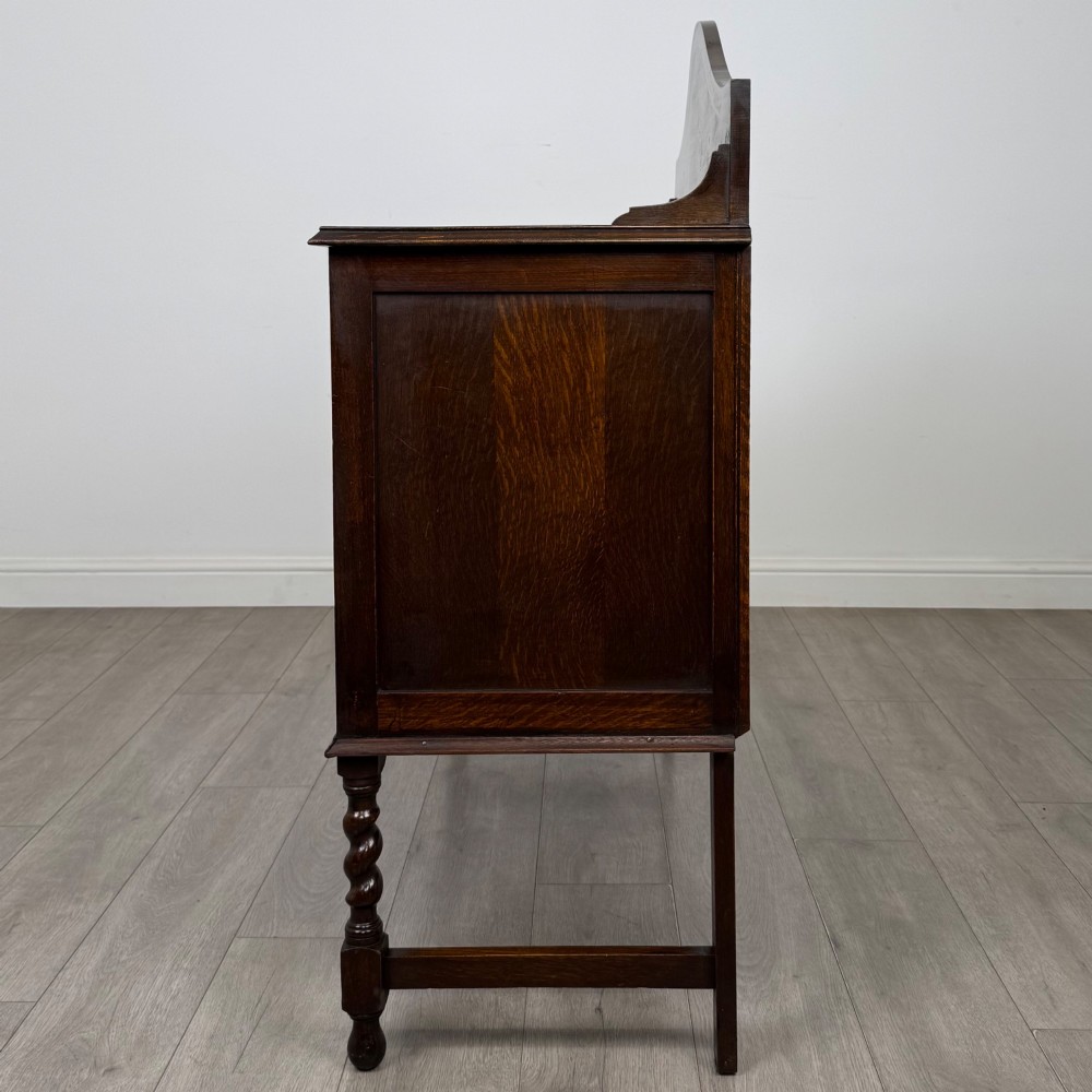 An Edwardian dark oak sideboard featuring a shaped upstand gallery, two top drawers with brass drop handles, twin raised-panel cupboard doors, and front barley twist legs. View IV