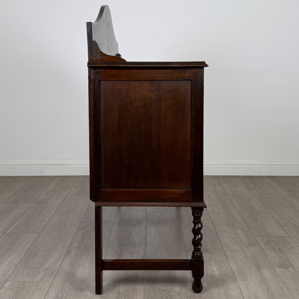 An Edwardian dark oak sideboard featuring a shaped upstand gallery, two top drawers with brass drop handles, twin raised-panel cupboard doors, and front barley twist legs. View VIII