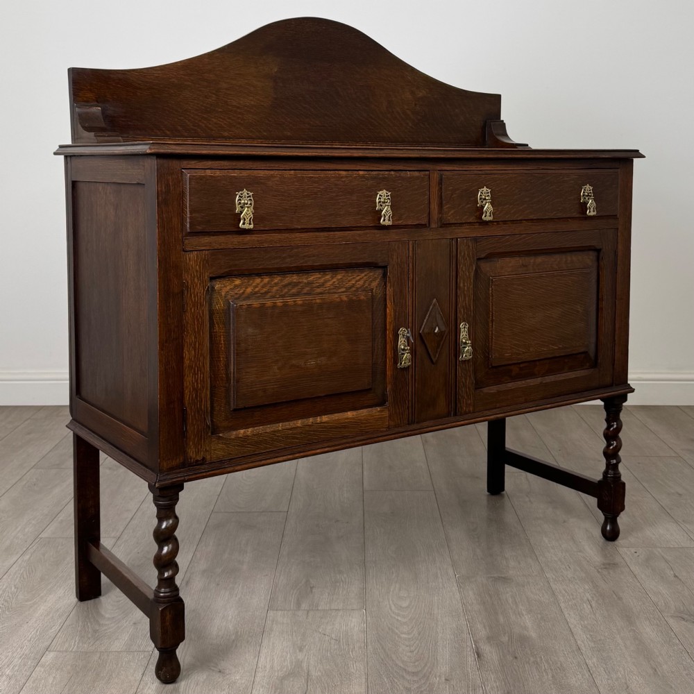 An Edwardian dark oak sideboard featuring a shaped upstand gallery, two top drawers with brass drop handles, twin raised-panel cupboard doors, and front barley twist legs. View IX