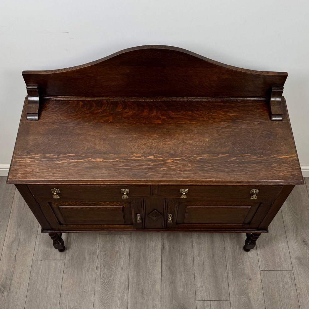 An Edwardian dark oak sideboard featuring a shaped upstand gallery, two top drawers with brass drop handles, twin raised-panel cupboard doors, and front barley twist legs. View X