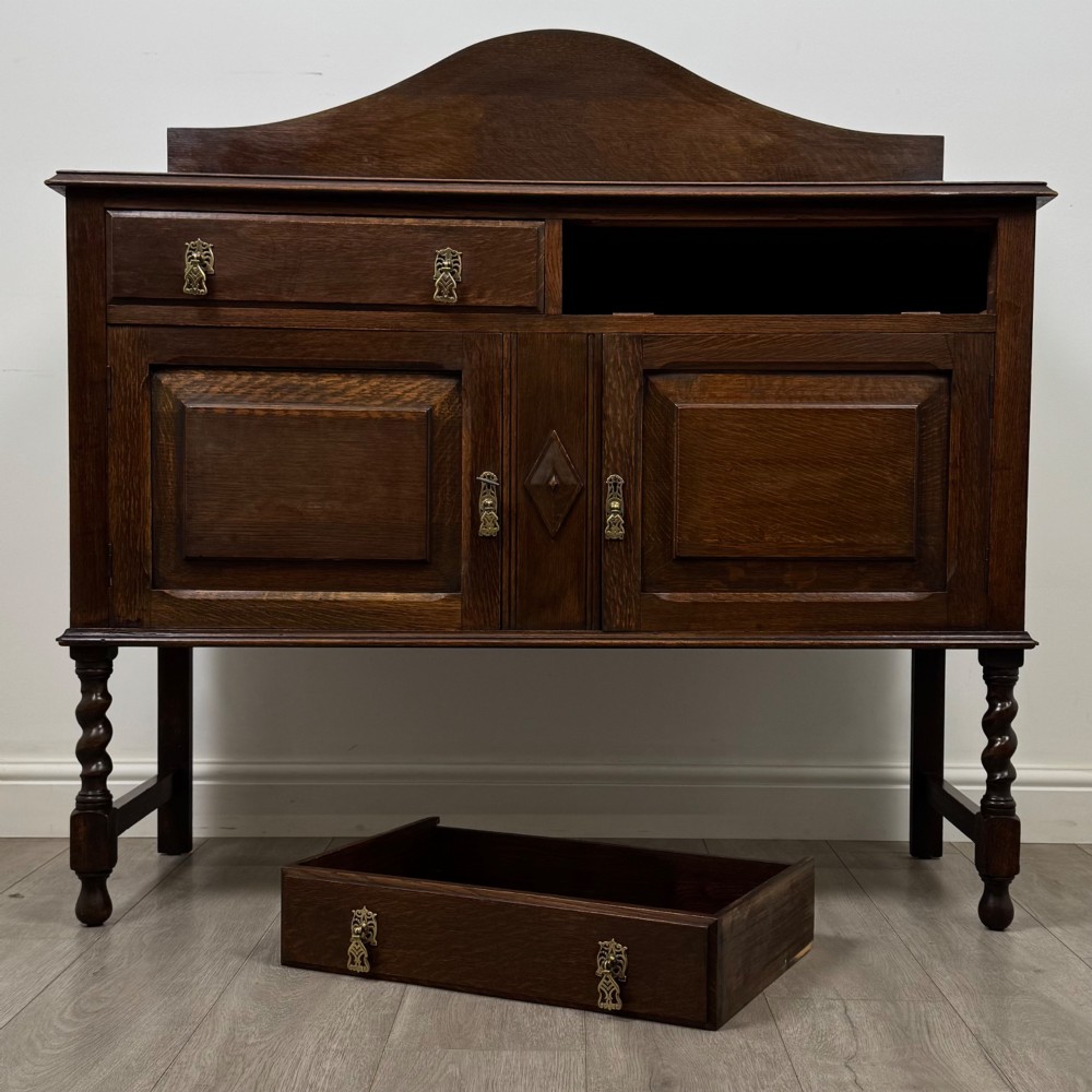 An Edwardian dark oak sideboard featuring a shaped upstand gallery, two top drawers with brass drop handles, twin raised-panel cupboard doors, and front barley twist legs. View XI