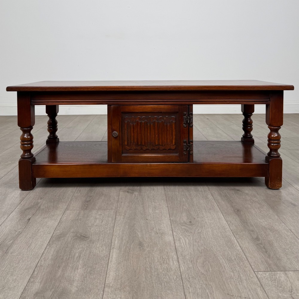 A low rectangular warm brown oak coffee table featuring turned baluster legs, a lower shelf, and a central cupboard door decorated with carved linenfold panelling.