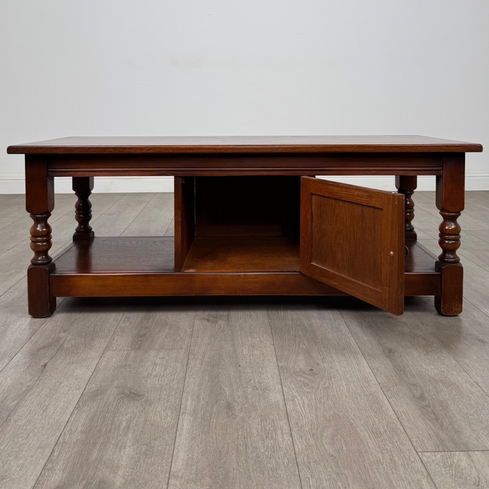 A low rectangular warm brown oak coffee table featuring turned baluster legs, a lower shelf, and a central cupboard door decorated with carved linenfold panelling. View II
