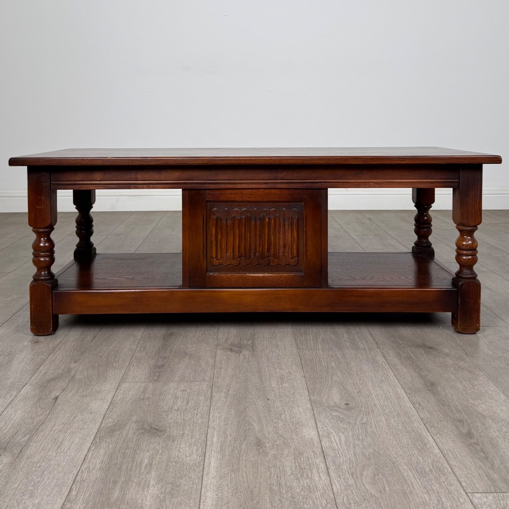 A low rectangular warm brown oak coffee table featuring turned baluster legs, a lower shelf, and a central cupboard door decorated with carved linenfold panelling. View VI