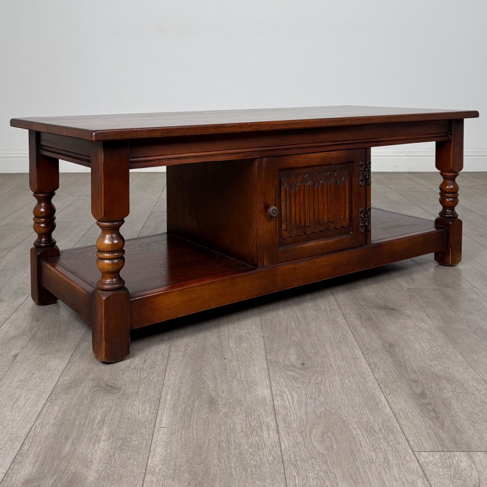 A low rectangular warm brown oak coffee table featuring turned baluster legs, a lower shelf, and a central cupboard door decorated with carved linenfold panelling. View IX