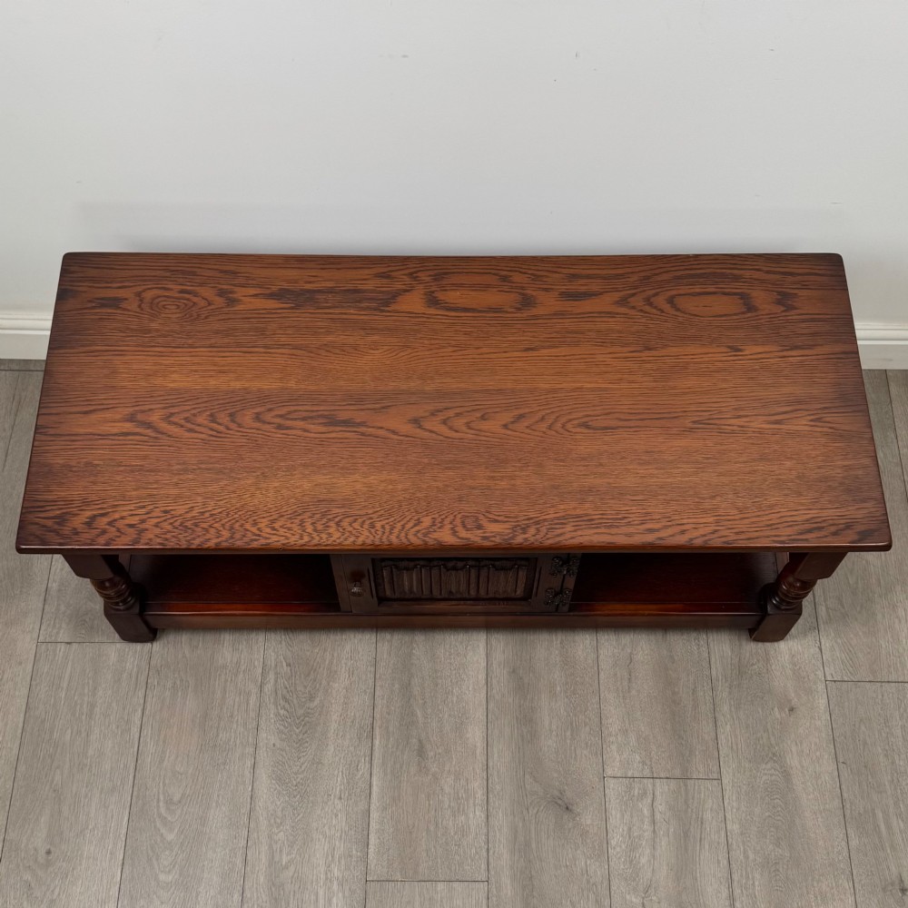 A low rectangular warm brown oak coffee table featuring turned baluster legs, a lower shelf, and a central cupboard door decorated with carved linenfold panelling. View X
