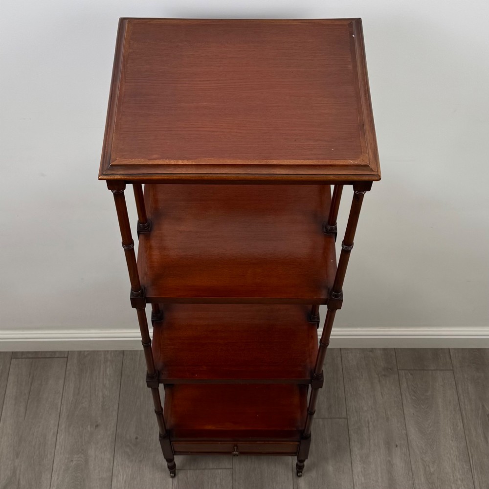 A tall, narrow 20th-century mahogany open bookcase featuring four tiered shelves supported by turned columns and a single drawer at the base raised on brass castors. View X