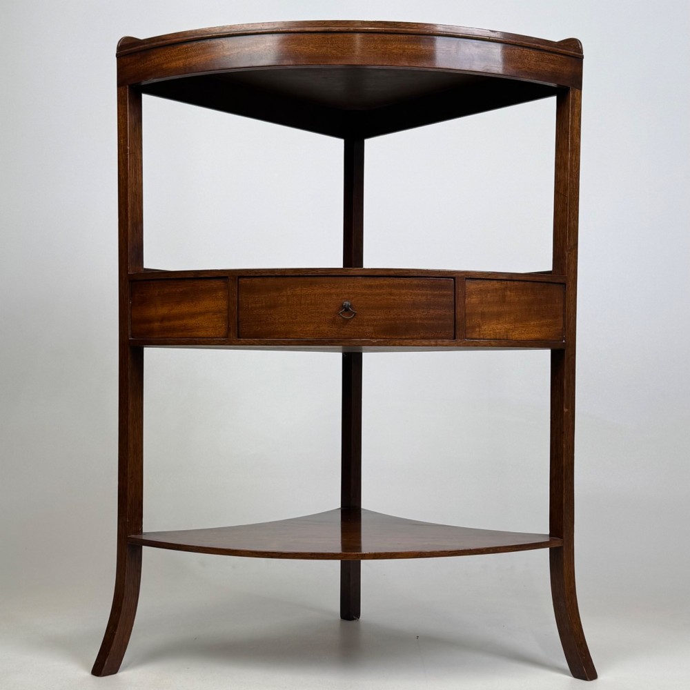A three-tiered Georgian style mahogany corner stand featuring a curved front, single drawer with brass ring pull, and splayed legs.