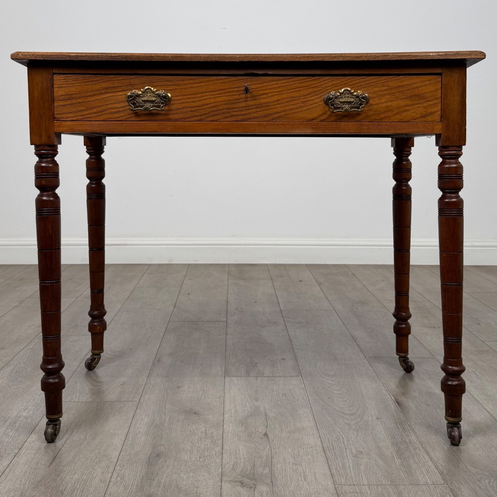 A 19th-century Victorian golden oak writing table featuring a single frieze drawer with decorative brass handles and ring-turned legs on castors.