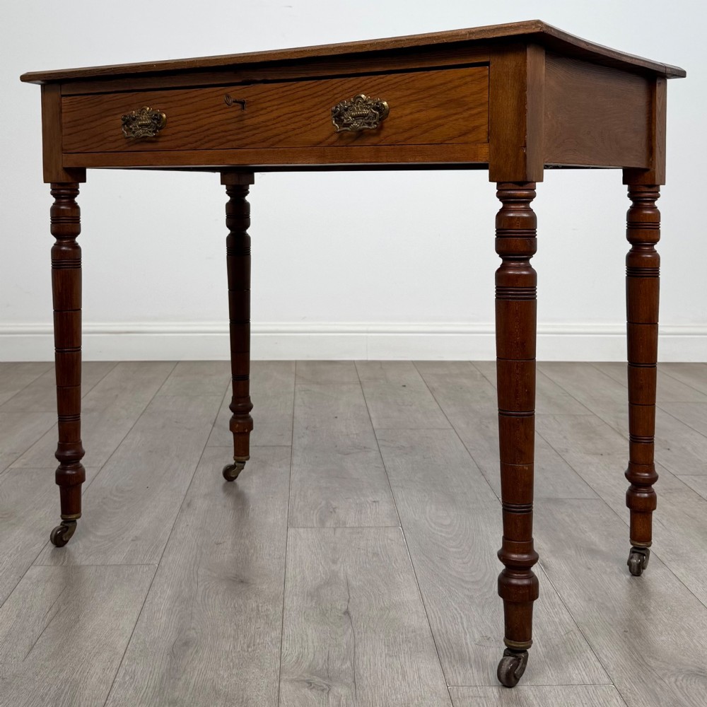 A 19th-century Victorian golden oak writing table featuring a single frieze drawer with decorative brass handles and ring-turned legs on castors. View II