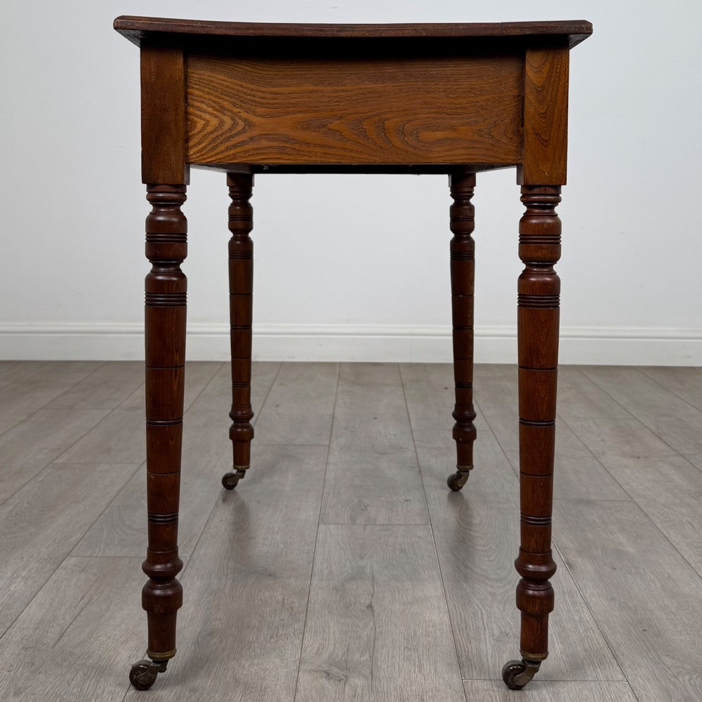 A 19th-century Victorian golden oak writing table featuring a single frieze drawer with decorative brass handles and ring-turned legs on castors. View III