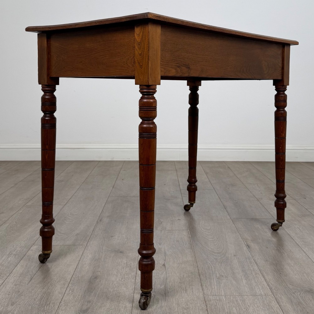 A 19th-century Victorian golden oak writing table featuring a single frieze drawer with decorative brass handles and ring-turned legs on castors. View IV