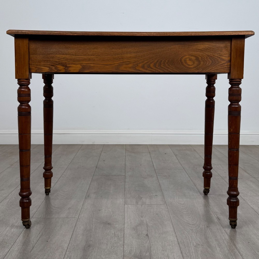 A 19th-century Victorian golden oak writing table featuring a single frieze drawer with decorative brass handles and ring-turned legs on castors. View V