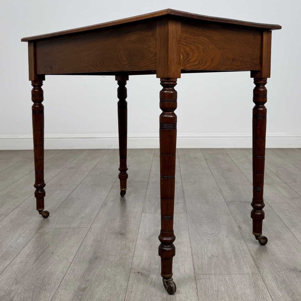 A 19th-century Victorian golden oak writing table featuring a single frieze drawer with decorative brass handles and ring-turned legs on castors. View VI