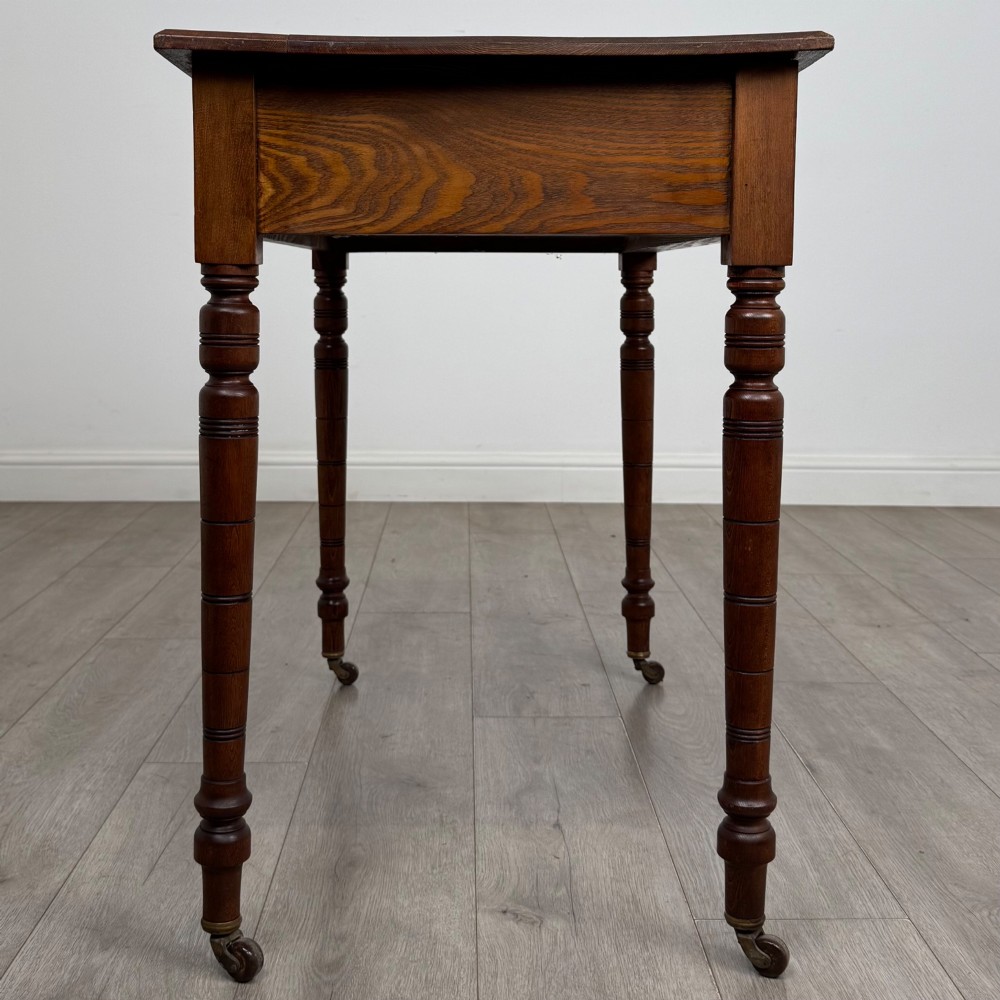 A 19th-century Victorian golden oak writing table featuring a single frieze drawer with decorative brass handles and ring-turned legs on castors. View VII
