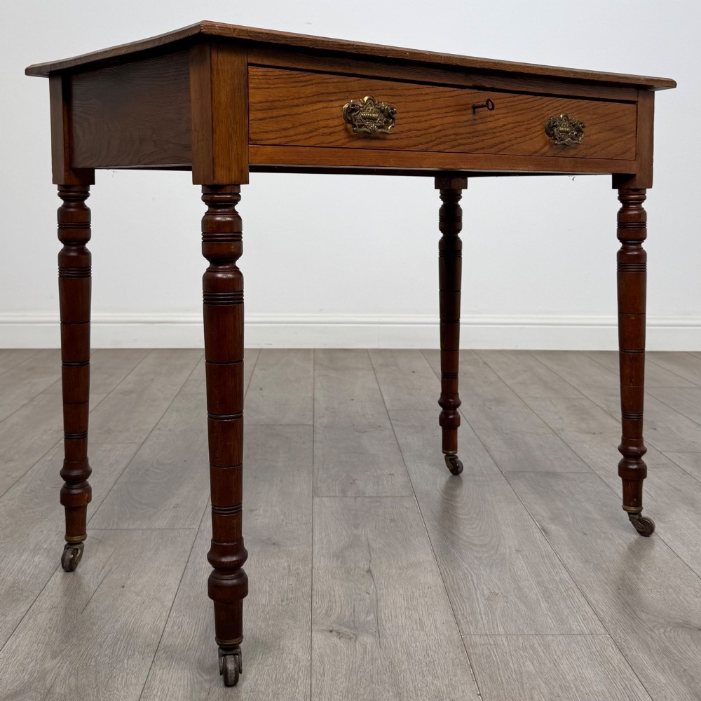 A 19th-century Victorian golden oak writing table featuring a single frieze drawer with decorative brass handles and ring-turned legs on castors. View VIII