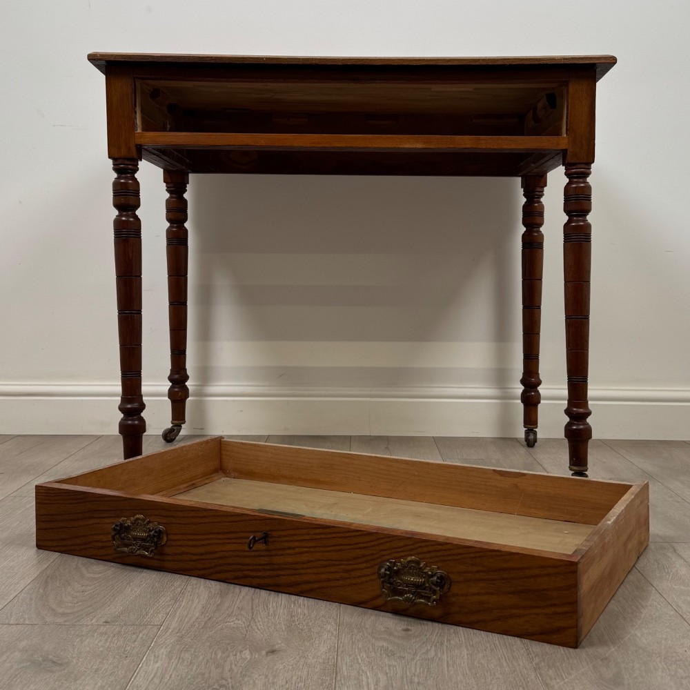 A 19th-century Victorian golden oak writing table featuring a single frieze drawer with decorative brass handles and ring-turned legs on castors. View X