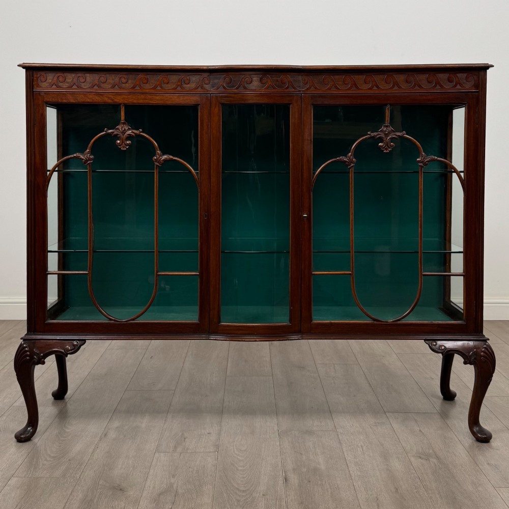 An early twentieth-century mahogany display cabinet featuring three glazed doors with curved astragal glazing bars, a carved top frieze, green fabric interior, and cabriole legs.
