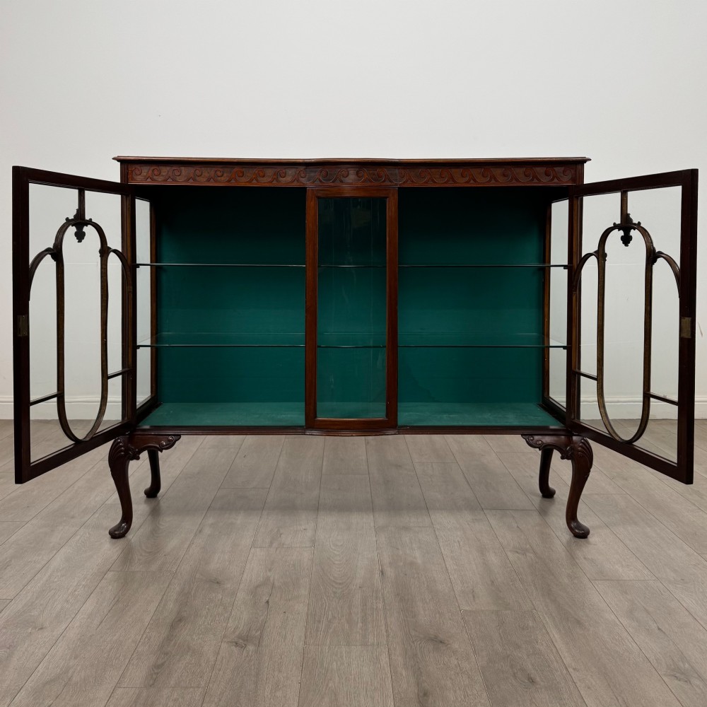 An early twentieth-century mahogany display cabinet featuring three glazed doors with curved astragal glazing bars, a carved top frieze, green fabric interior, and cabriole legs. View II